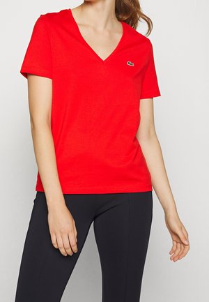 Vrouw draagt een felrode T-shirt met V-hals en korte mouwen, met een klein groen krokodillenlogo, gecombineerd met zwarte nauwsluitende broek, tegen een effen achtergrond.