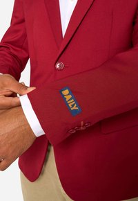 Man die de mouw van een rood colbert met een gele en blauwe "DAILY" patch op de manchette rechtzet, draagt een wit overhemd en beige broek.