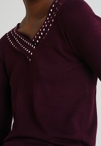 Burgundy knit top with a V-neck and white polka dot accents along the neckline. Fitted long sleeves, soft texture, and minimal design.