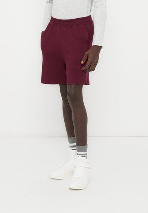 Uomo con pantaloncini bordeaux a vita elasticizzata, maglia a maniche lunghe grigio chiaro, calzini grigi a righe e sneakers bianche alte, in piedi su sfondo bianco.