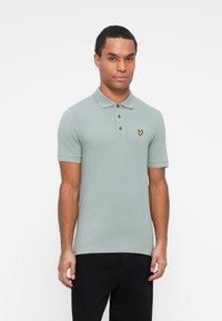 Gai zaļas kokvilnas polo krekls ar klasisku apkakli, trīs pogu aizdari un mazu dzeltenu logo kreisajā krūtī. Melni bikses.