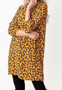 Gul tunika klänning med svart blommönster, 3/4-ärmar, lös passform, rundad halsringning och sidofickor, i kombination med svarta leggings.