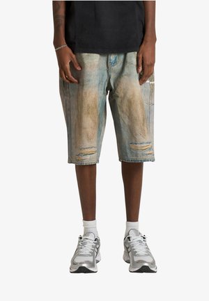 Persona che indossa pantaloni corti lunghi in denim sbiaditi e consumati, calzini bianchi e scarpe da ginnastica sportive argentate, in piedi con le mani rilassate lungo i fianchi.