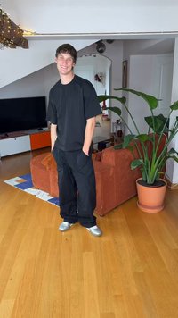 Black oversized t-shirt, loose black pants, and silver sneakers. Indoor setting with a brown corduroy sofa and large green plant nearby.