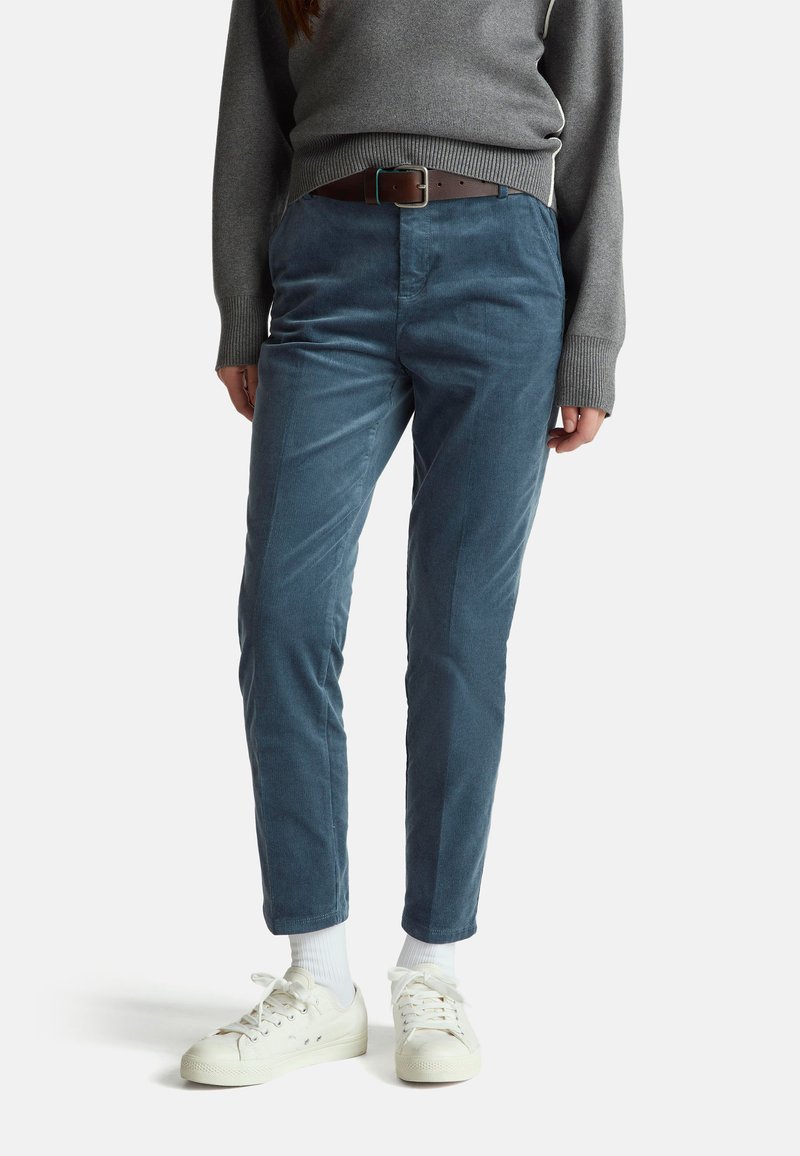 Cordhosen in einem sanften Blauton, mit einem klassischen Schnitt und subtiler Hochglanz. Kombiniert mit einem grauen Pullover und weißen Turnschuhen.
