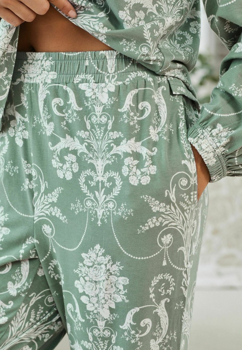 Zöld szövet loungewear szett, amely bonyolult fehér virágmintákkal díszített, rugalmas derékpánttal és oldalsó zsebekkel a praktikusság és kényelem érdekében.