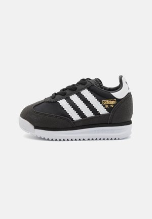 Zapatillas deportivas en negro y gris con rayas blancas, parte superior de ante y malla, suela de goma y detalles de marca en dorado en el lateral.