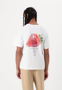 Witte katoenen T-shirt met een grote, gedetailleerde aardbeienafbeelding op de achterkant, samen met recepttekst eronder in zwarte letters.