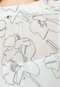 Gros plan d’un tissu blanc transparent avec des dessins floraux linéaires noirs et une petite étiquette à triple bandes à la couture du col d’un vêtement.