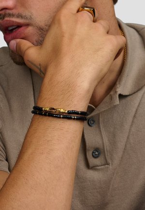 Sort perlearmbånd med guld accenter på et håndled, prydet med en blanding af runde perler og en dekorativ lås, sat mod en neutral stofbaggrund.