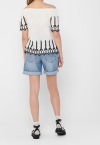 Haut blanc épaules dénudées avec accents brodés noirs et manches courtes, assorti à un short en denim retroussé. Sandales noires à brides.