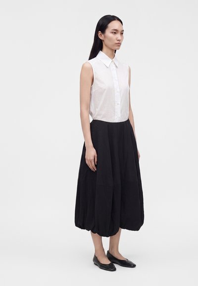 Femme debout portant une chemise blanche sans manches boutonnée, une jupe mi-longue noire volumineuse et des chaussures plates noires sur un fond uni.