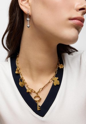 Zbliżenie kobiety noszącej złoty naszyjnik z zawieszkami w kształcie klucza, serca, kokardki i misia oraz srebrną kolczyk w kształcie strzały.