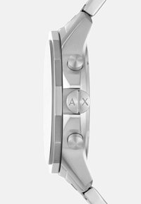 Hõbedatooniline käekell, millel on ümmargune korpus, metallist rihm, kaks külg-nuppu ja tekstuuriga pind, millel on üksiku AX logo ühe nupu peal.
