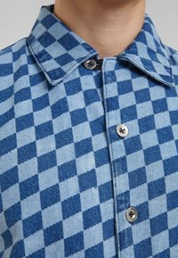 Camicia a quadri blu e azzurri in materiale denim. Presenta un colletto, chiusura con bottoni e cuciture dettagliate sul tessuto.