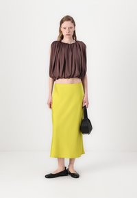 Brown balloon-sleeve crop top, bright yellow midi skirt, and black flat shoes. Model holds a small black bag. Simple, smooth textures.