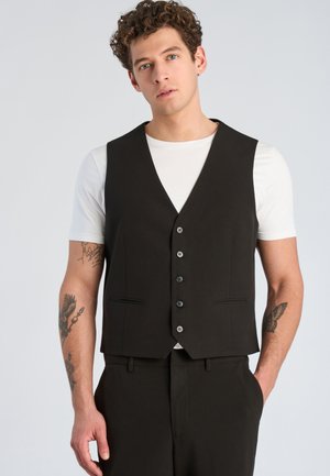 Junger Mann mit lockigem Haar, trägt eine schwarze Knopfweste über einem weißen T-Shirt, mit Tätowierungen auf beiden Unterarmen, steht vor einem einfarbigen Hintergrund.