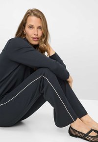 Zwart loungewear-set bestaande uit een langärmelig shirt en wijde broek met witte bies aan de zijkanten, gecombineerd met zwarte, gevlochten platte schoenen.