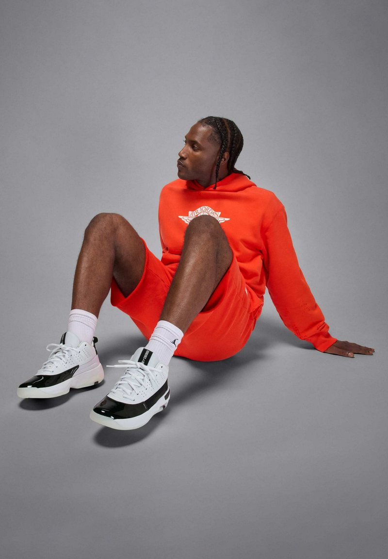 Mand i rød Air Jordan hoodie og shorts siddende på gulvet med bøjede ben, iført hvide og sorte sneakers samt hvide strømper.
