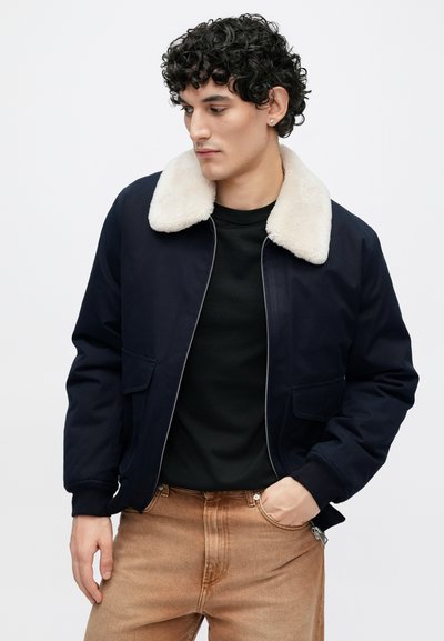 Mornarska bomber jakna s bež ovratnikom, ki spominja na krzno, sprednjimi žepi in zadrgo. Kombinirana s črno majico in rjavimi hlačami.