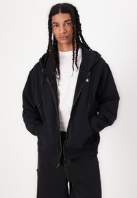 Schwarze Zip-Hoodie mit großer Kapuze, Fronttaschen und einem weißen Logo auf der Brust. Getragen über einem weißen T-Shirt und kombiniert mit dunklen Hosen.
