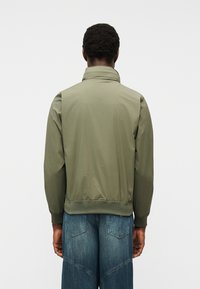 Άνδρας με κοντά σγουρά μαλλιά, φοράει μια ελιάς πράσινη ζακέτα και μπλε τζιν παντελόνι, απεικονίζεται από πίσω με φόντο απλό.