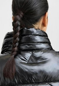 Doudoune noire avec un col haut, texture brillante et patch de marque triangulaire. Cheveux tressés. Vue de dos.