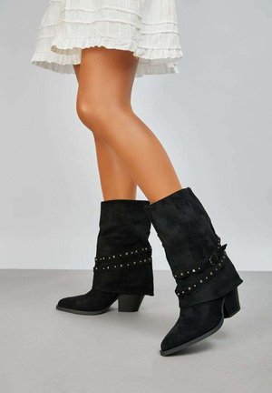 Femme portant des bottes mi-mollet en suède noir avec des sangles cloutées et une jupe blanche à volants, debout devant un fond gris clair uni.