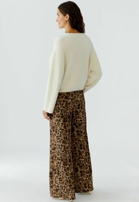 Vrouw die naar links kijkt, gekleed in een wit, fuzzy, langwerpig top met lange mouwen en brede leopardprintbroek, tegen een effen lichte achtergrond.