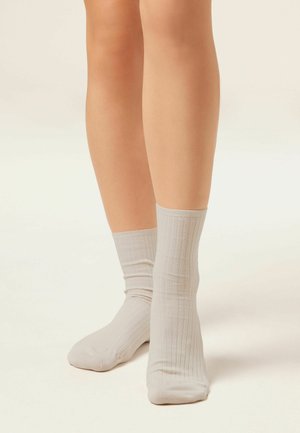 Gambe con calzini a coste color beige chiaro alti fino al polpaccio, in piedi su una superficie neutra con uno sfondo chiaro.