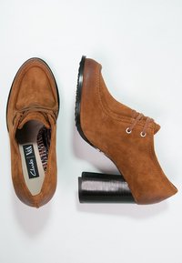 Zapatos de tacón de ante marrón con punta redondeada, dos ojales para los cordones y un tacón de bloque negro texturizado. El forro presenta un diseño estampado.