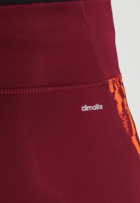 Bordowe legginsy sportowe z teksturowanym pomarańczowym wzorem po boku, z białym logo "climalite" oraz gładkim, elastycznym materiałem.