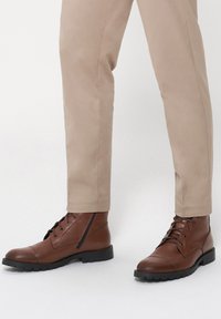 Brązowe skórzane botki sięgające do kostki, z sznurowaniem, zaokrąglonym noskiem i teksturowaną boczną panelem. Połączone z beżowymi zwężanymi spodniami. Czarna gumowa podeszwa.