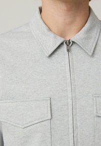 Lys grå zip-up jakke med et tekstureret mønster, krave og to brystlommer. Har en metallynlås og en ren finish.