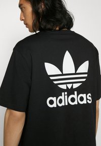 Čierne bavlnené tričko s veľkým bielym logom Adidas trefoil, troch pruhov a slovom "adidas" písmom tučným bielym písmom.