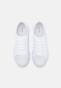 Zapatillas de lona blancas con punta redondeada, seis ojales, cordones planos blancos y suela de goma. El interior presenta un nombre de marca impreso.