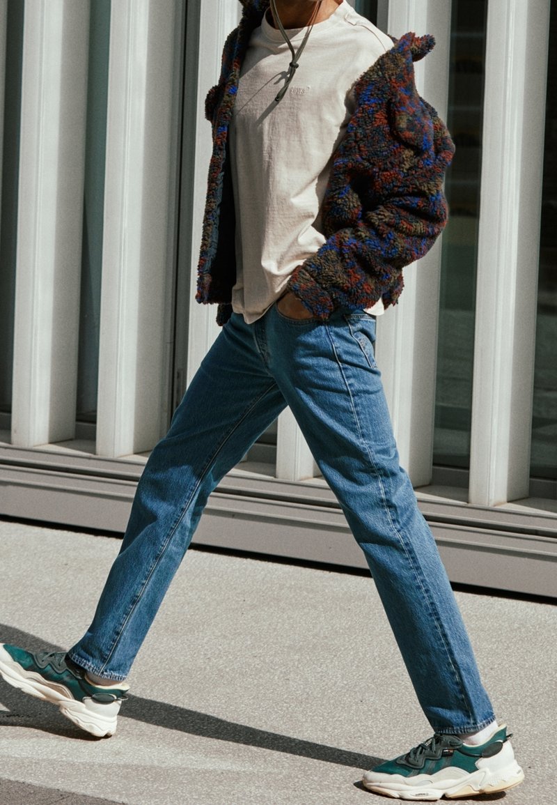 Casual outfit featuring a fuzzy, multicolored jacket, a cream T-shirt, blue jeans, and chunky green and white sneakers.