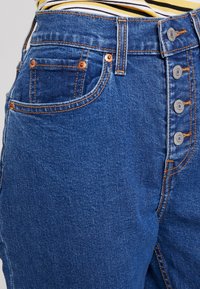 Džínové kalhoty s vysokým pasem, vybavené pěti knoflíky, modrou látkou, oranžovým stehem a boční kapsou s měděnými kovovými doplňky.