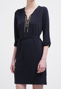 Mörkblå klänning med V-ringning, trekvartsärmar, guldkedjedetaljer och ett midjeband. Slät tyg med en avslappnad passform.