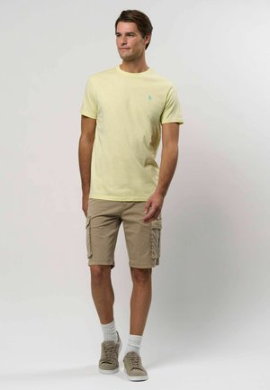 Svetlo žlté tričko s okrúhlym výstrihom, skombinované s béžovými cargo šortkami s bočnými vreckami. Šedé tenisky a biele ponožky po členky dotvárajú vzhľad.