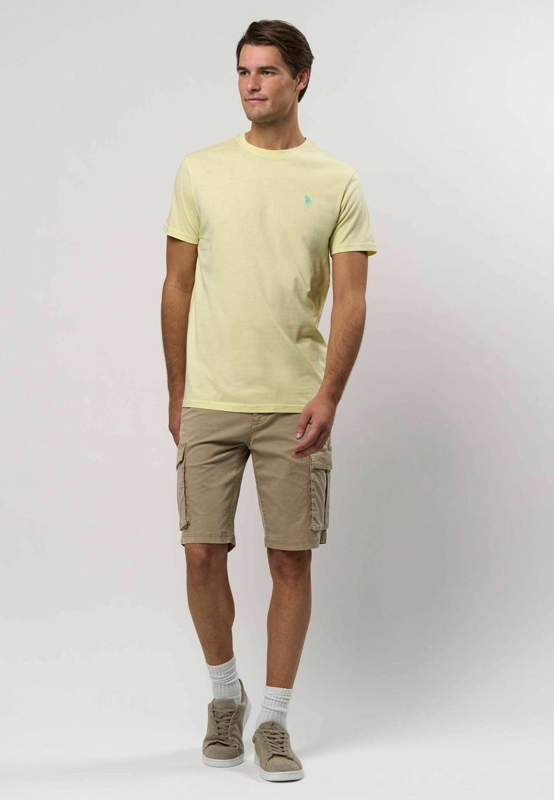 Šviesiai geltonas apvalios iškirptės marškinėliai, derinami su smėlio spalvos cargo šortais su šoninėmis kišenėmis. Pilki sportbačiai ir balti sportiniai kojinės papildo įvaizdį.