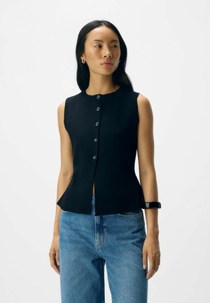 Top nero senza maniche con scollatura alta e chiusura a bottone, caratterizzato da un corpetto aderente che si allarga in vita, indossato con jeans azzurri chiari.