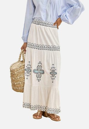 Mujer con una falda larga blanca con patrones bordados en negro y azul, blusa a rayas azul claro, llevando un bolso tejido de paja y sandalias color beige.