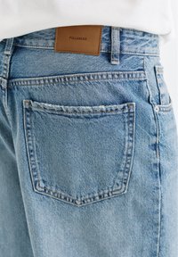 Jean en denim bleu clair avec une poche arrière et une étiquette de marque en cuir marron indiquant "PULL&BEAR" à la taille.