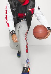 Chaqueta varsity en blanco y negro, sudadera con capucha roja, pantalones de chándal grises con el estampado "CHICAGO", zapatillas coloridas, sosteniendo un balón de baloncesto.