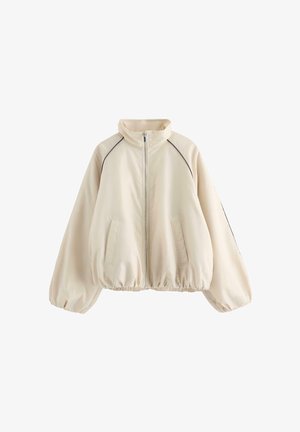 Beige jakke med lynlås, høj krave, elastisk kant og ærmegab, to forlommer samt sort piping langs skuldersømmene.