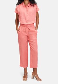 Nainen, joka pukeutunut korallinväriseen lyhythihaiseen napitettuun puseroon ja siihen sopiviin naruilla kiristettäviin housuihin, yhdistettynä beigeihin korkeakorkoisiin sandaaleihin, seisoo käsine taskuissa.