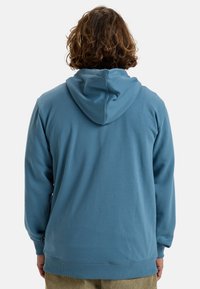 Sweatshirt à capuche bleu clair avec une coupe décontractée, des poignets côtelés et un détail de poche kangourou. Texture lisse et design minimaliste.