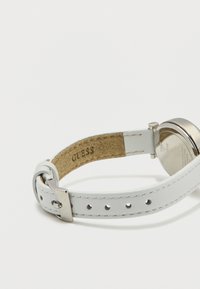 Λουράκι ρολογιού από λευκό δέρμα με μεταλλική αγκράφα, μερικώς κουμπωμένο, που εμφανίζει το όνομα της μάρκας "GUESS" στην εσωτερική πλευρά. Οπίσθιο μέρος θήκης ρολογιού από ασημί μέταλλο ορατό.