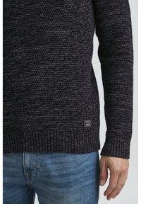 Dunkler Strickpullover mit einem strukturierten Muster, gerippten Bündchen und Saum sowie einem kleinen Etikett am unteren Saum, kombiniert mit hellblauen Jeans.